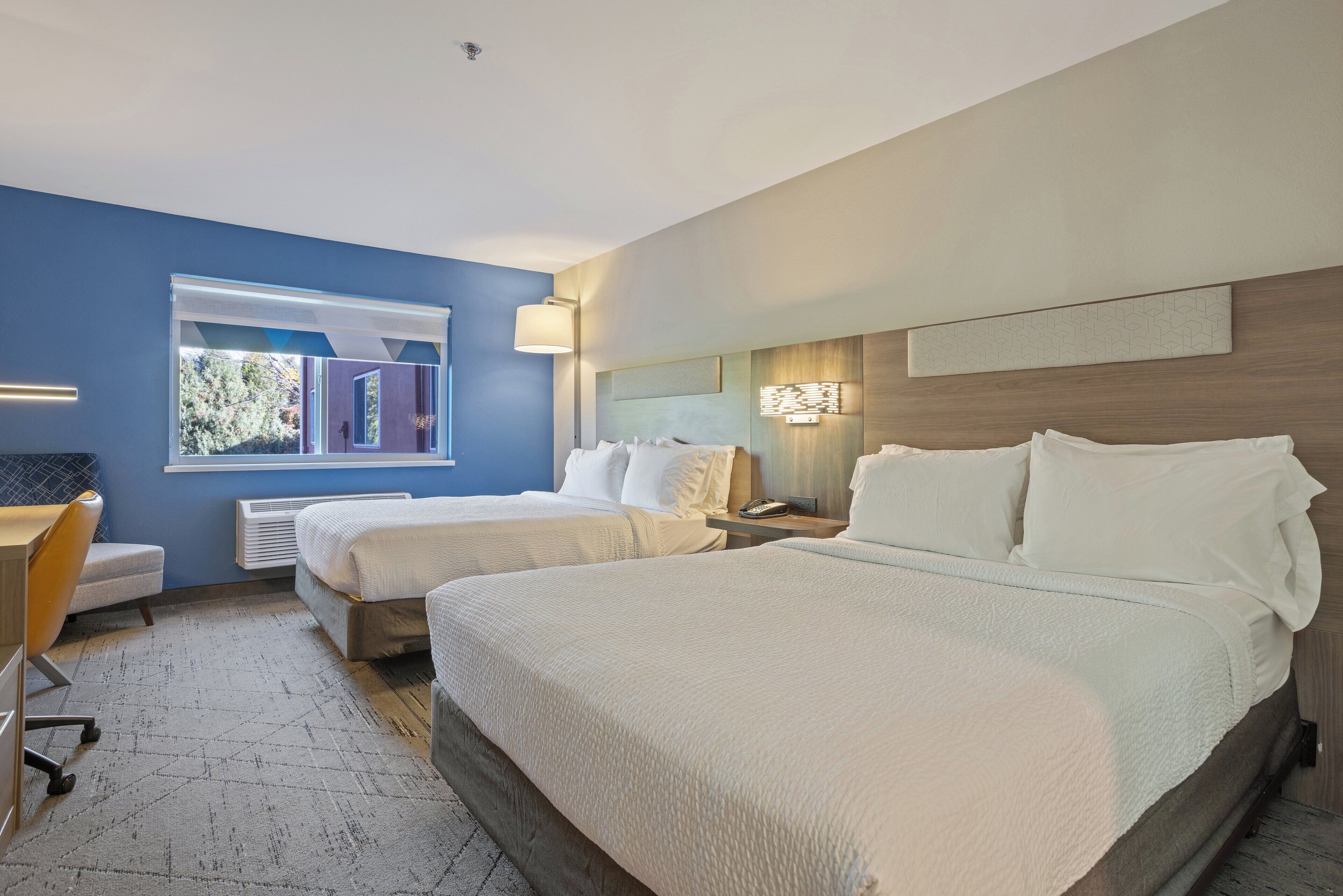 A hotel room with two beds, a desk, and a window with a view of greenery.