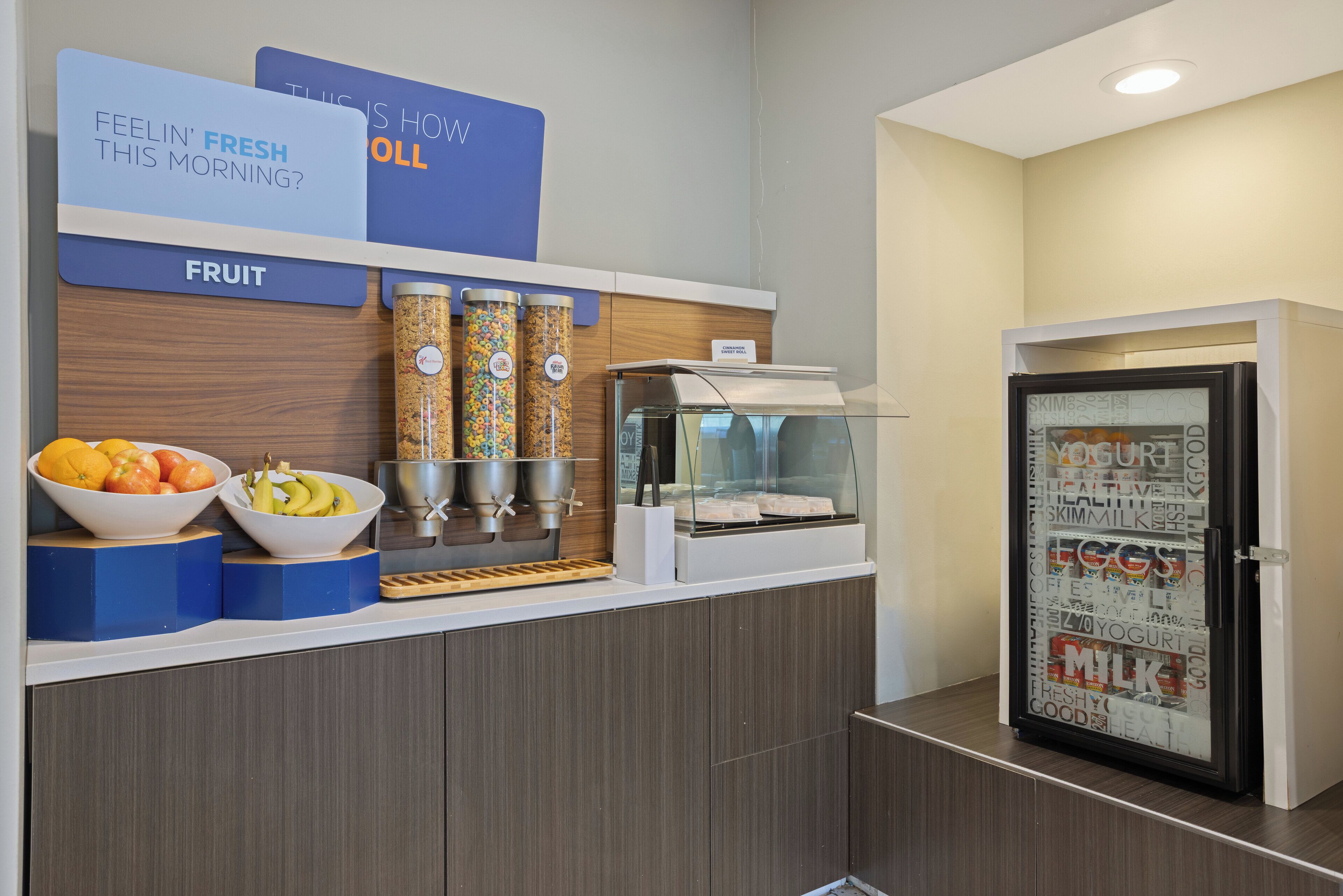 A breakfast bar with fruit bowls, a coffee machine, and a refrigerator with a milk dispenser.