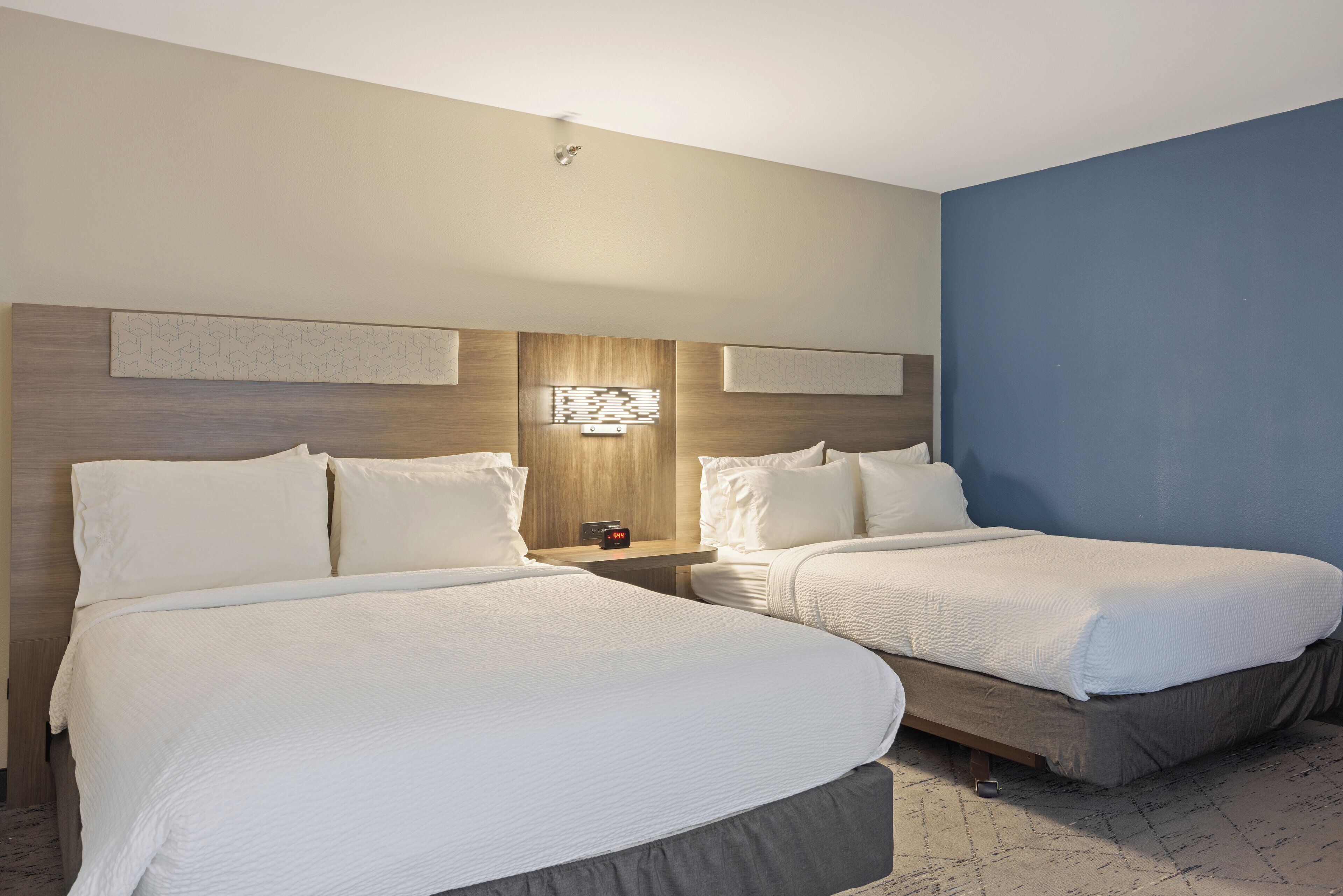 A hotel room with two beds, a nightstand with a clock, and a wall-mounted light fixture.