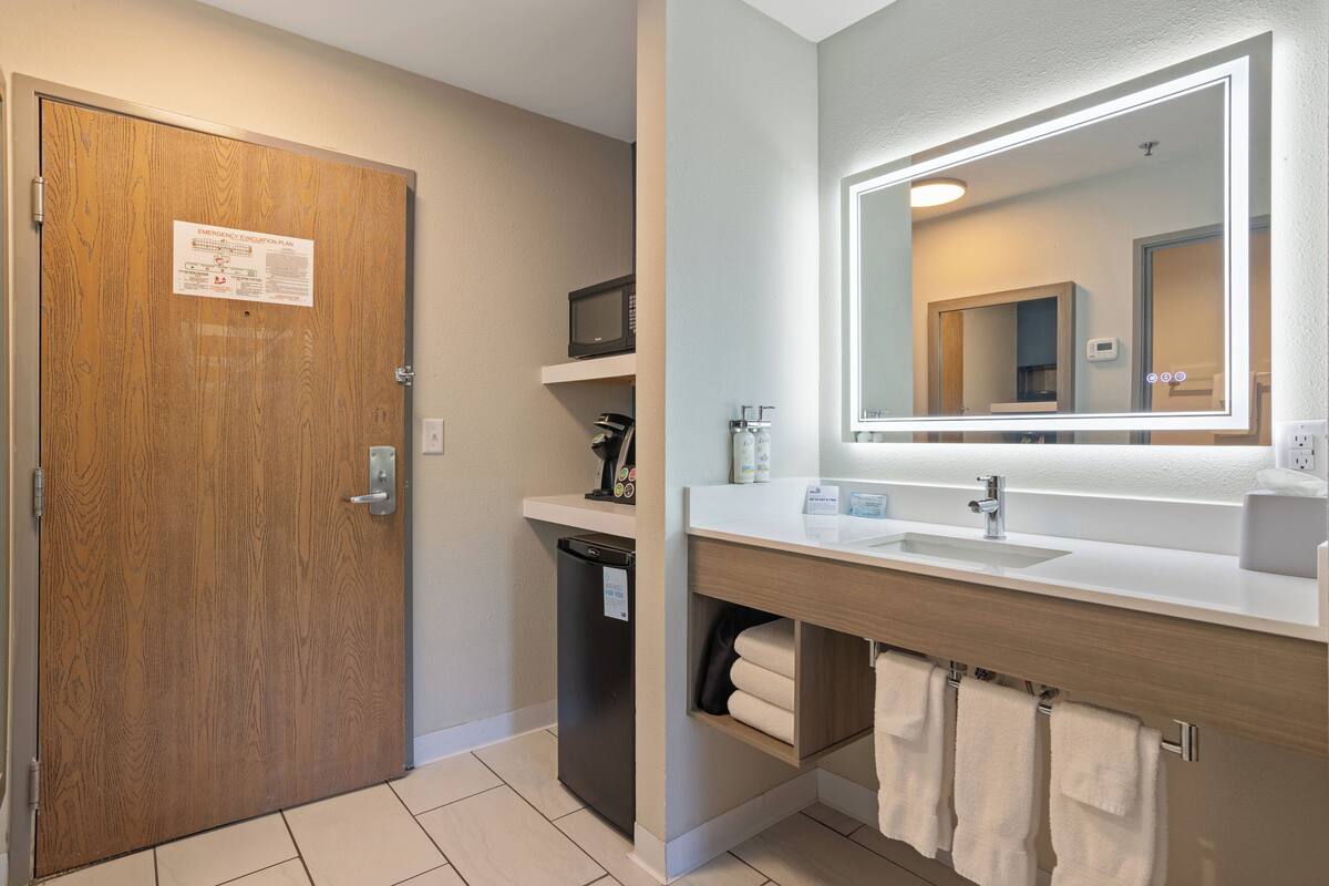 A bathroom with a wooden door, a microwave, a sink with a mirror, and towels.