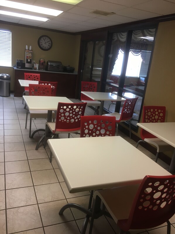 Interior de un restaurante con mesas blancas y sillas rojas, un reloj en la pared y un cubo de basura.