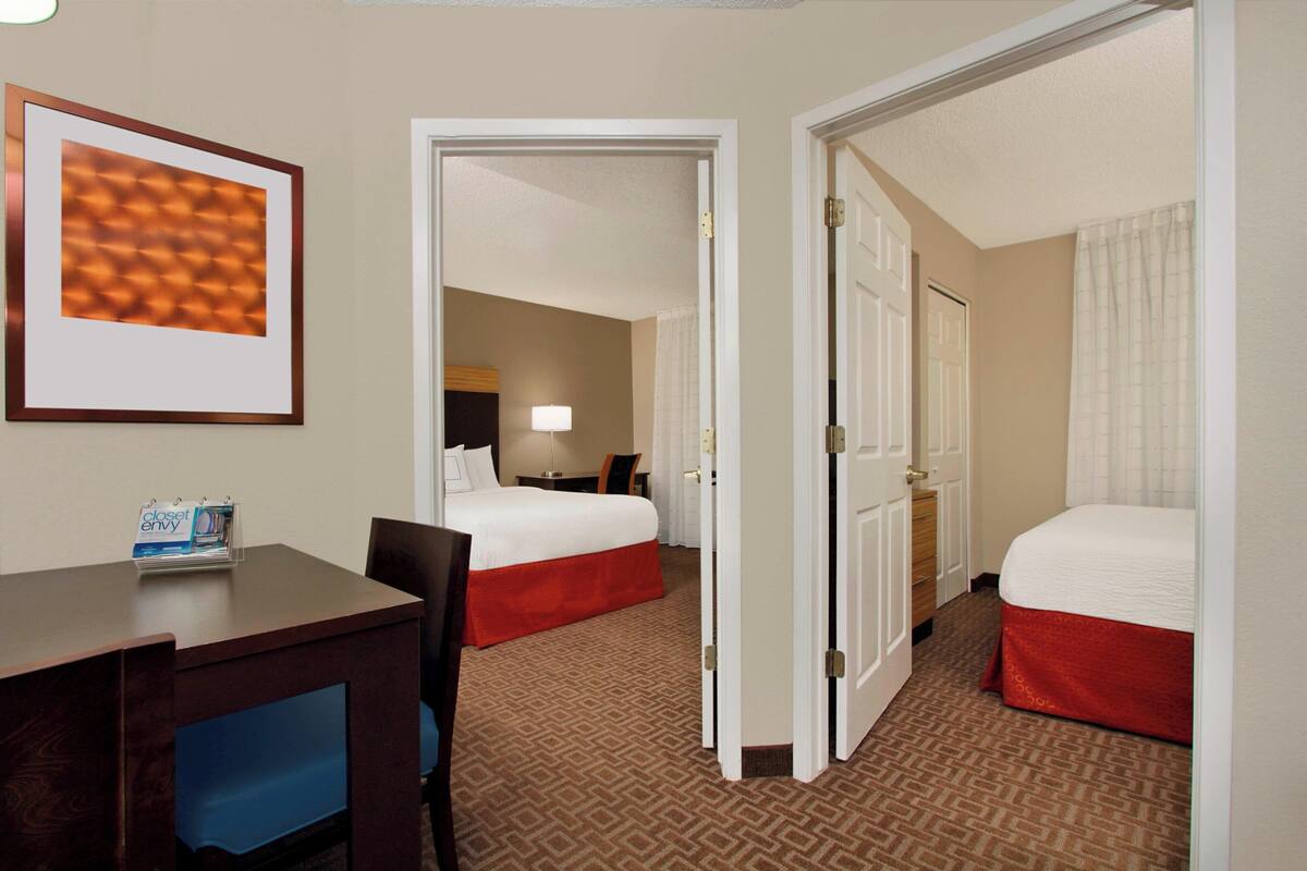 Une chambre d’hôtel avec deux lits, un bureau et une chaise. Il y a une porte qui donne sur une autre pièce et un tableau encadré accroché au mur.