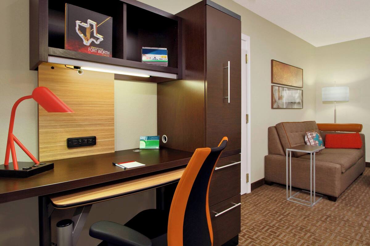 Une chambre d’hôtel avec un bureau en bois, une lampe rouge, un fauteuil de bureau noir, un canapé marron et une petite table avec une lampe.