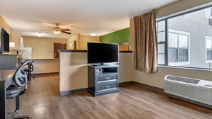 A hotel room with a flat-screen TV on a stand, a desk with a chair, a treadmill, a large window with curtains, and a ceiling fan.