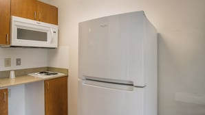 A kitchen with a white microwave, a white refrigerator, wooden cabinets, a granite countertop, and a stainless steel sink. - HomeTowne Studios By Red Roof Denver - Airport/ Aurora (Aurora)