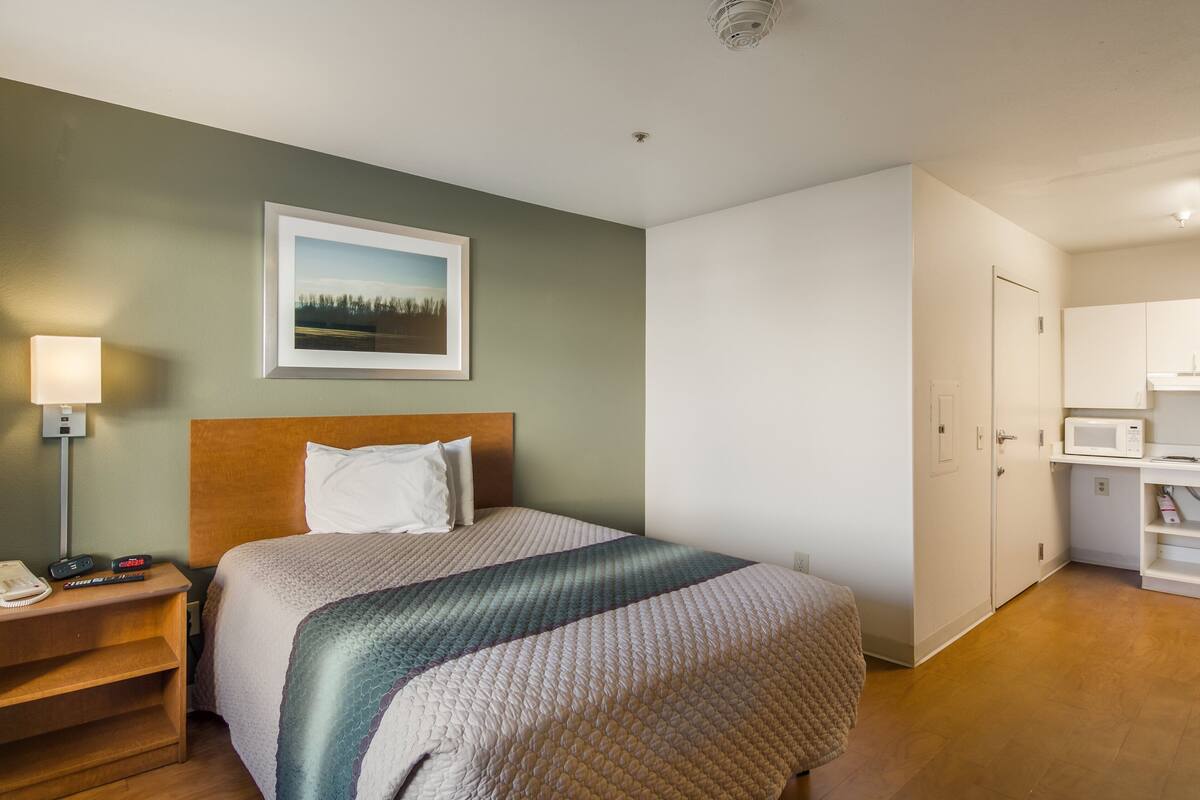 Ett hotellrum med en säng, ett nattduksbord, en lampa, en inramad tavla på väggen och en liten köksdel.