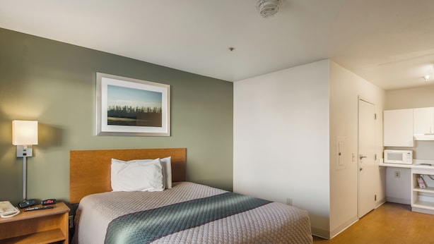 A hotel room with a bed, a nightstand, a lamp, a framed picture on the wall, and a kitchenette area. - HomeTowne Studios By Red Roof Denver - Airport/ Aurora (Aurora)