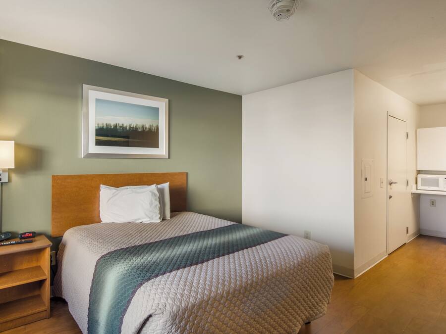 Quarto de hotel com cama, mesa de cabeceira, luminária, quadro pendurado na parede e área de cozinha compacta.