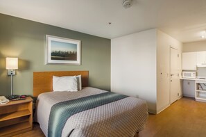 A hotel room with a bed, a nightstand, a lamp, a framed picture on the wall, and a kitchenette area. - HomeTowne Studios By Red Roof Denver - Airport/ Aurora (Aurora)