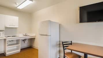 Una kitchenette compacta con un refrigerador blanco, un microondas y un lavamanos. Hay una mesa de madera con una silla y un televisor de pantalla plana montado en la pared.