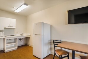 Una kitchenette compacta con un refrigerador blanco, un microondas y un lavamanos. Hay una mesa de madera con una silla y un televisor de pantalla plana montado en la pared.