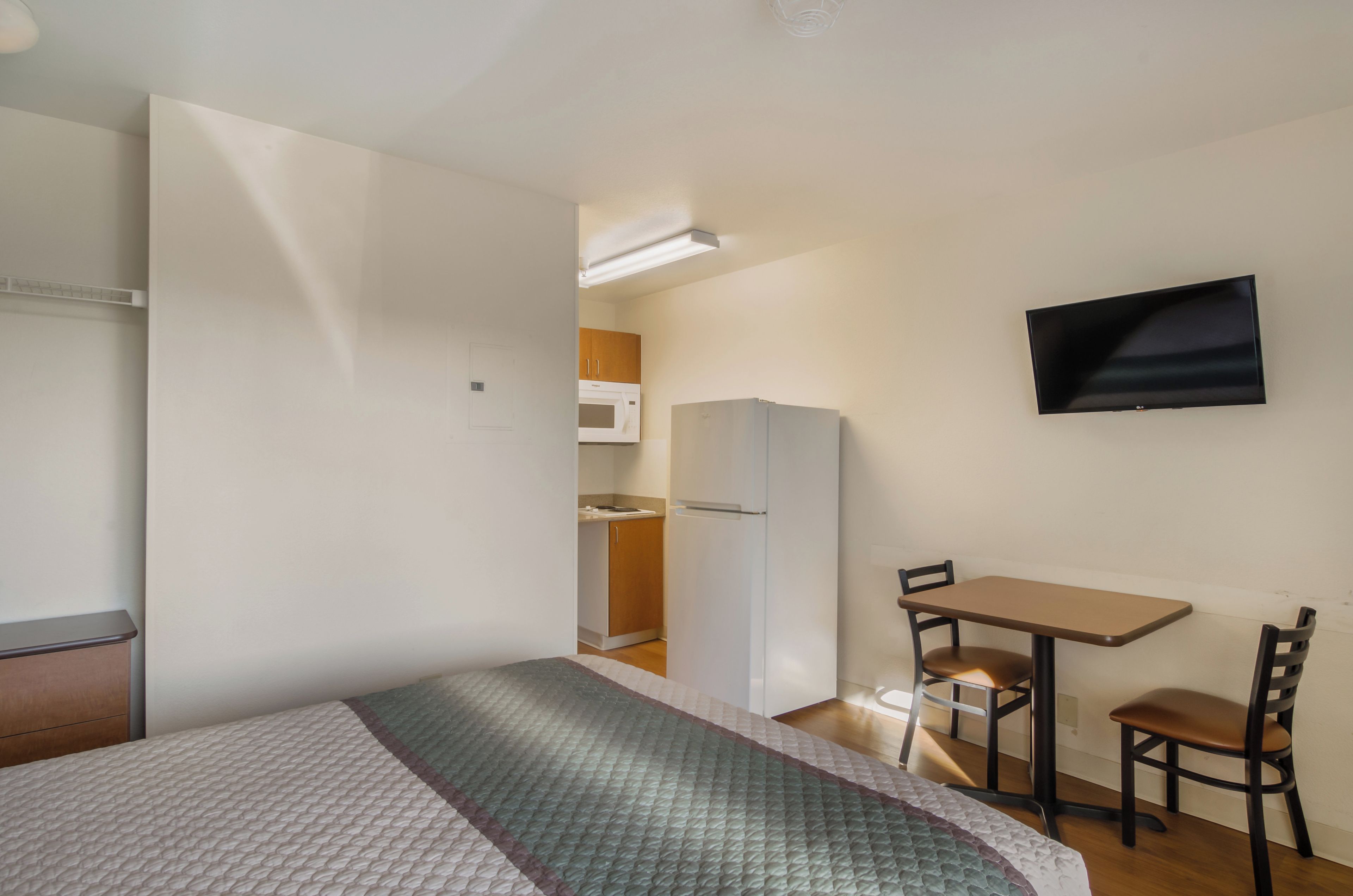 A compact room with a bed, a small table, two chairs, a television mounted on the wall, and a kitchenette in the background.