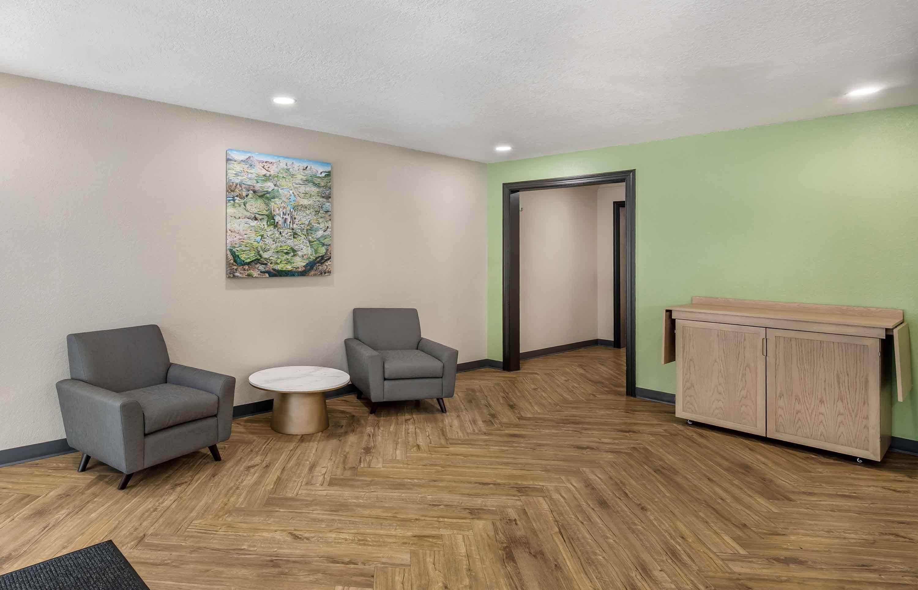 A reception area with two grey armchairs, a small round table, a wooden cabinet, and a painting on the wall.