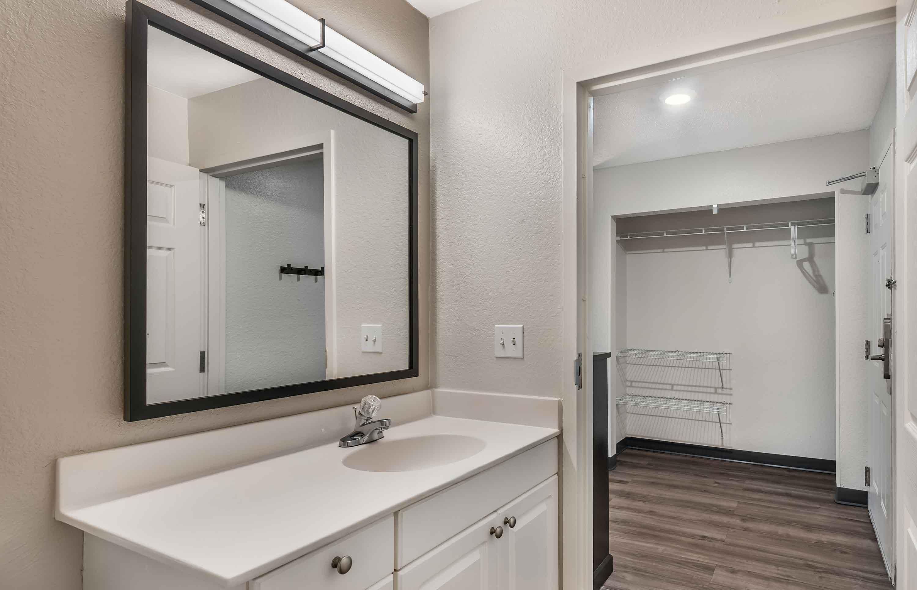 A bathroom with a white vanity, a large mirror, and a walk-in closet with a metal rack.