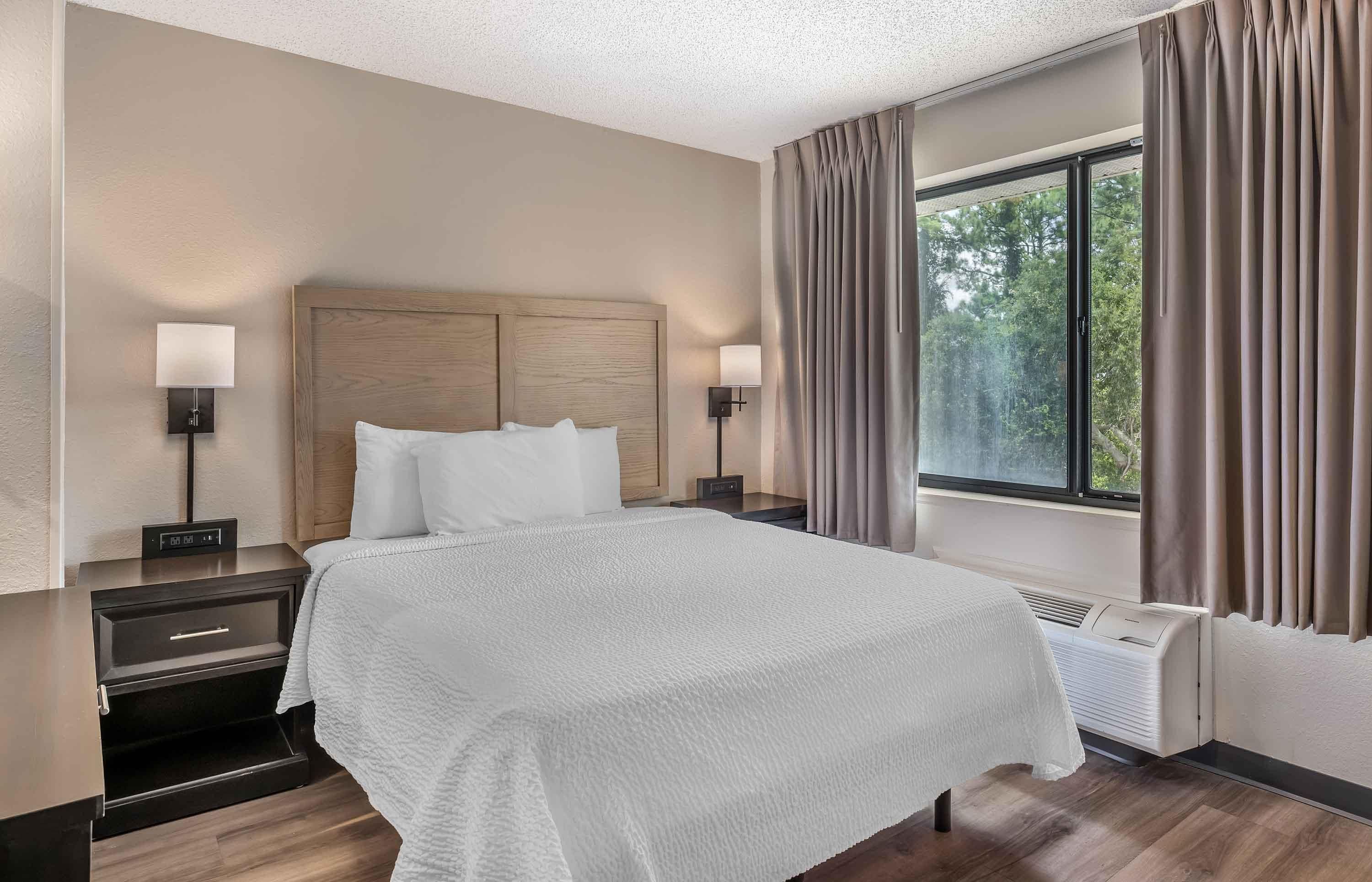 A hotel room with a large bed, two bedside tables with lamps, a window with curtains, and a wall-mounted heater.