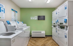 A laundry facility with multiple washing machines and dryers, a green wall, and informational signs. - Extended Stay America Suites Daytona Beach Speedway (Daytona Beach)
