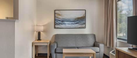 A modern living room with a grey sofa, a small wooden table, a lamp, and a painting on the wall.