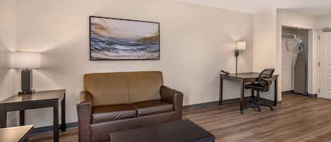 A hotel room with a brown leather sofa, a small table, a desk with a chair, a painting on the wall, and a coat rack with a coat.