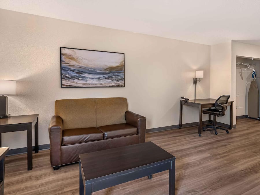 A hotel room with a brown leather sofa, a small table, a desk with a chair, a painting on the wall, and a coat rack with a coat.
