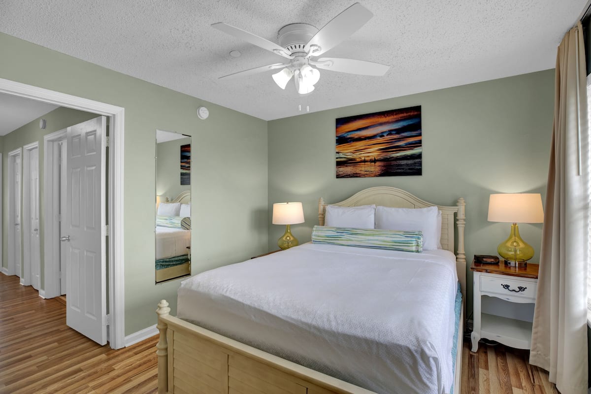 A hotel room with a bed, bedside lamps, a ceiling fan, a painting, and a mirror.