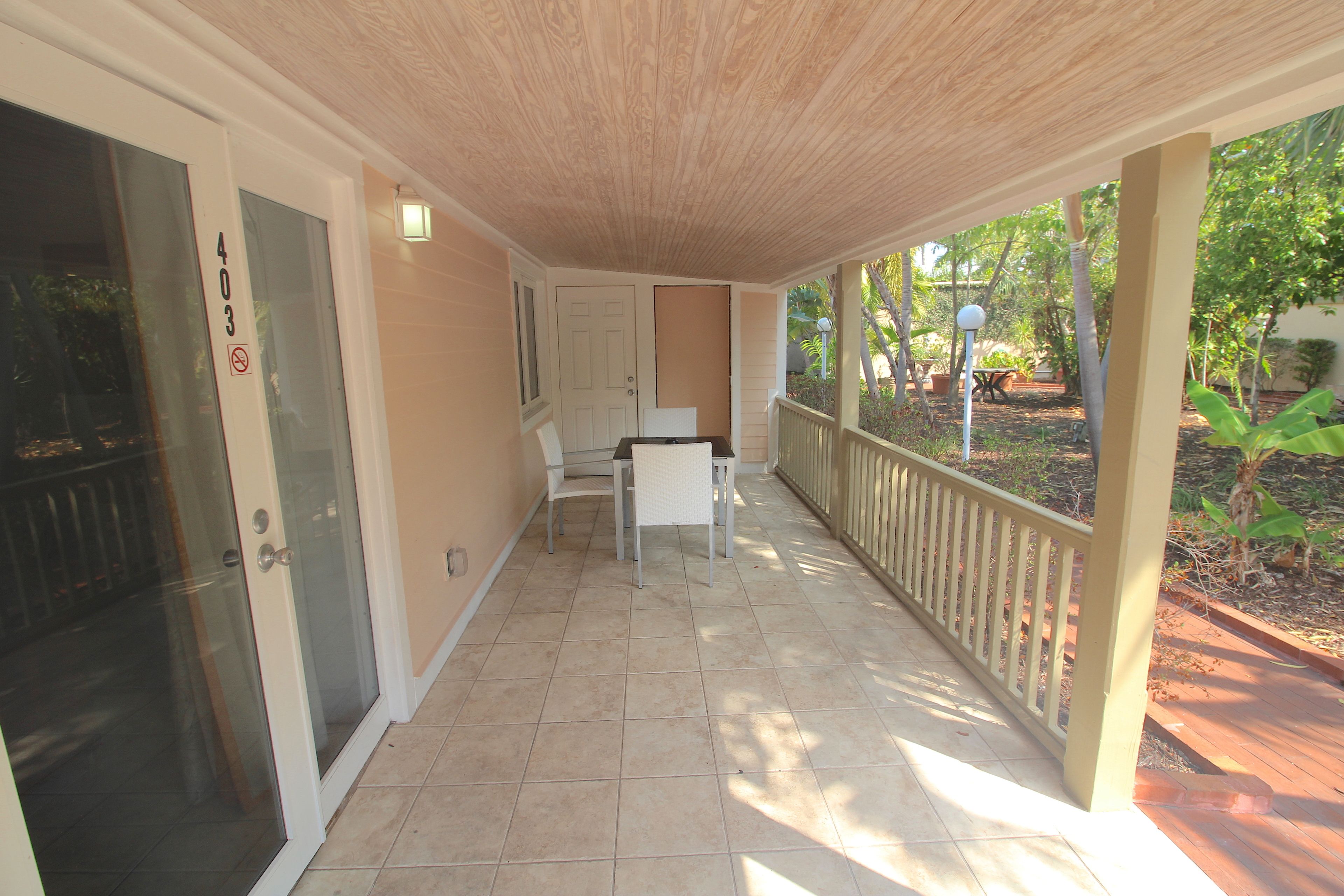 A covered porch with a white door, a small table and chair, and a railing overlooking a garden.
