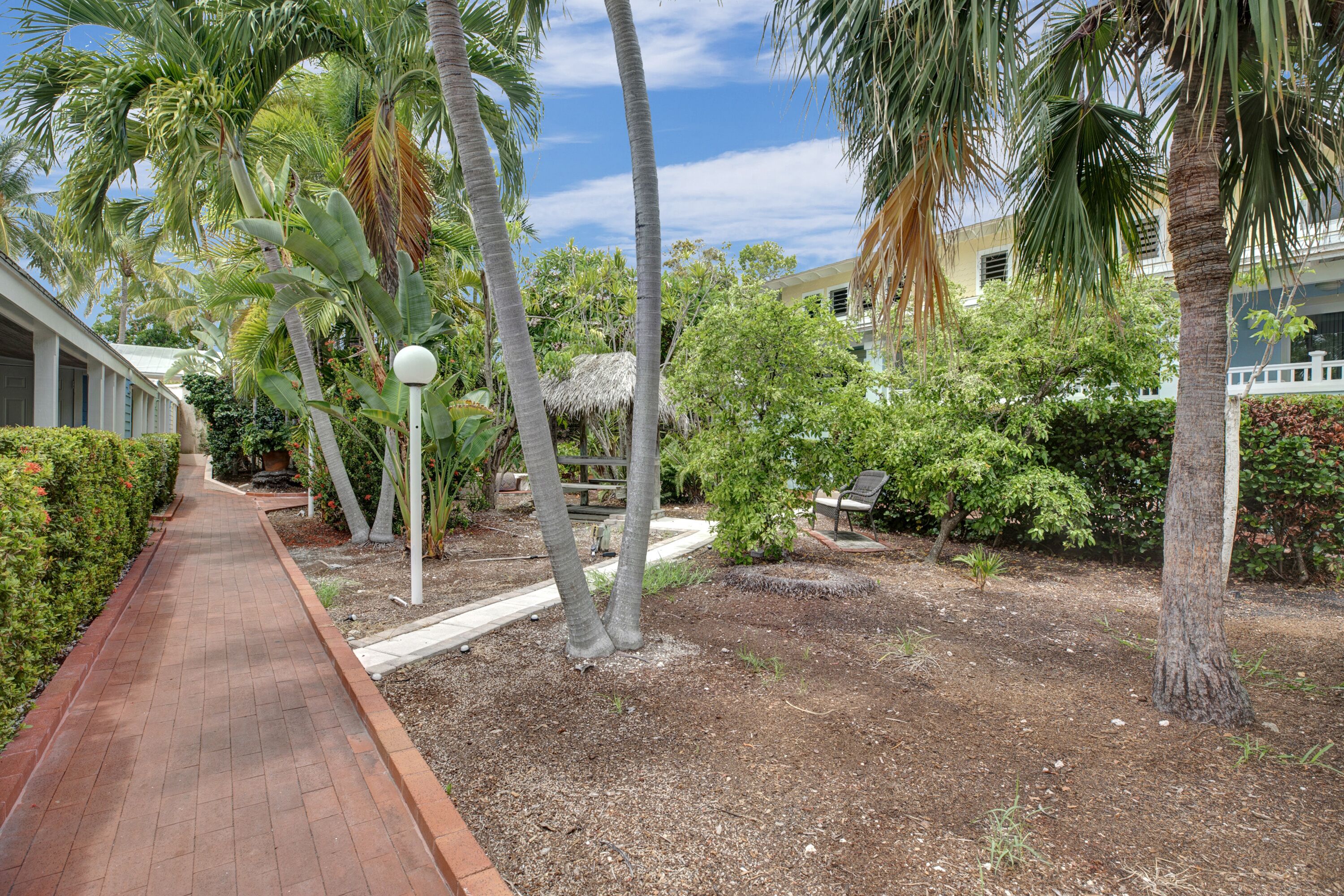 A paved pathway leading through a landscaped garden with palm trees and a thatched-roof structure.
