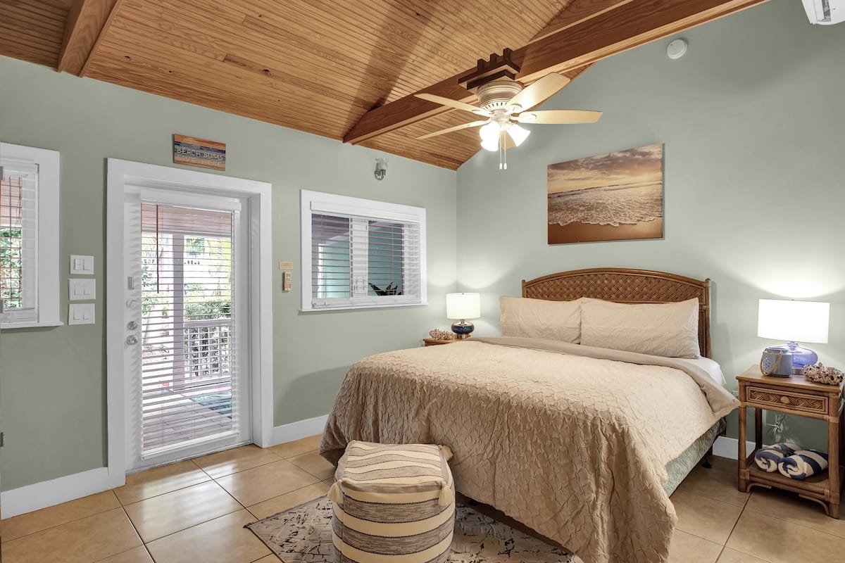 A bedroom with a wooden ceiling, a bed with beige bedding, a nightstand with a lamp, and a window with blinds.