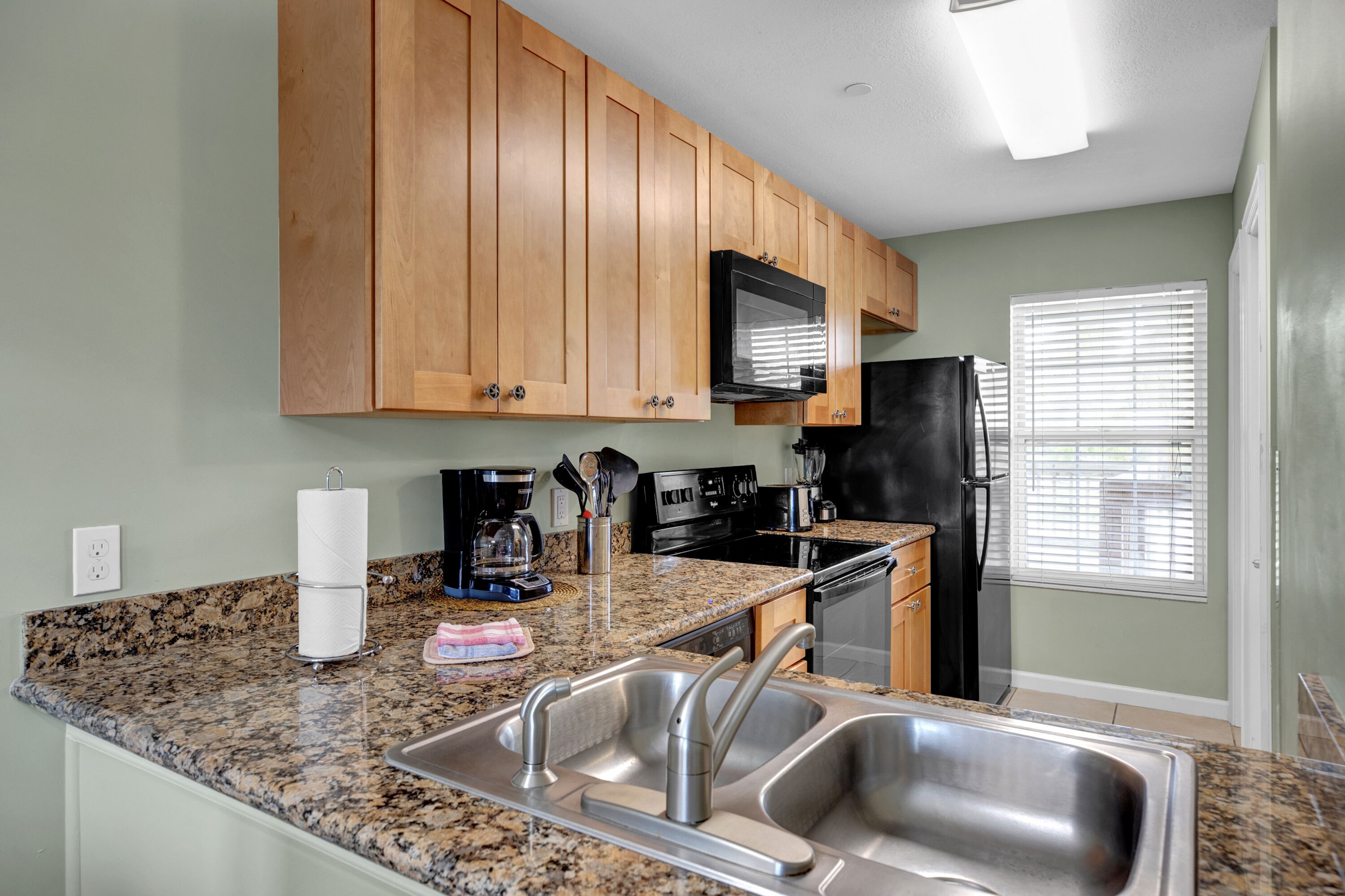 A kitchen with granite countertops, stainless steel sinks, wooden cabinets, a black refrigerator, and a microwave.