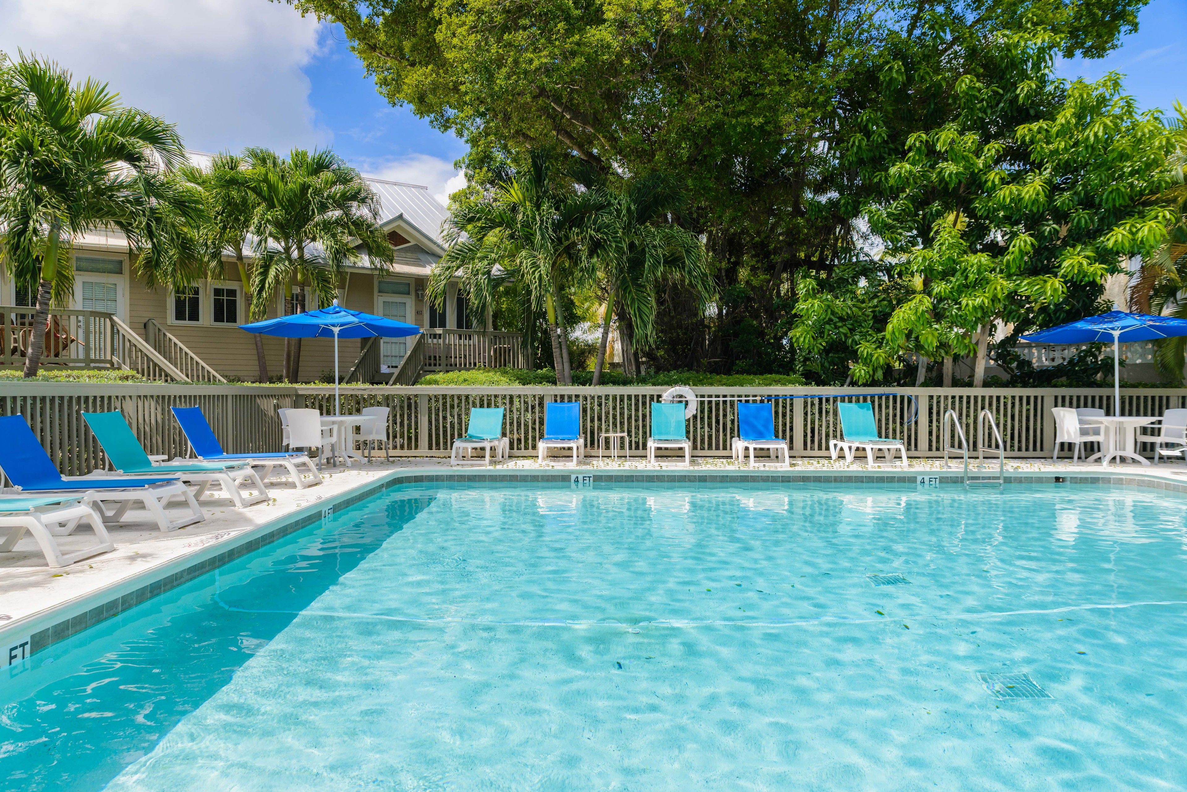 A swimming pool with blue chairs and umbrellas, surrounded by palm trees and a building with a covered deck.