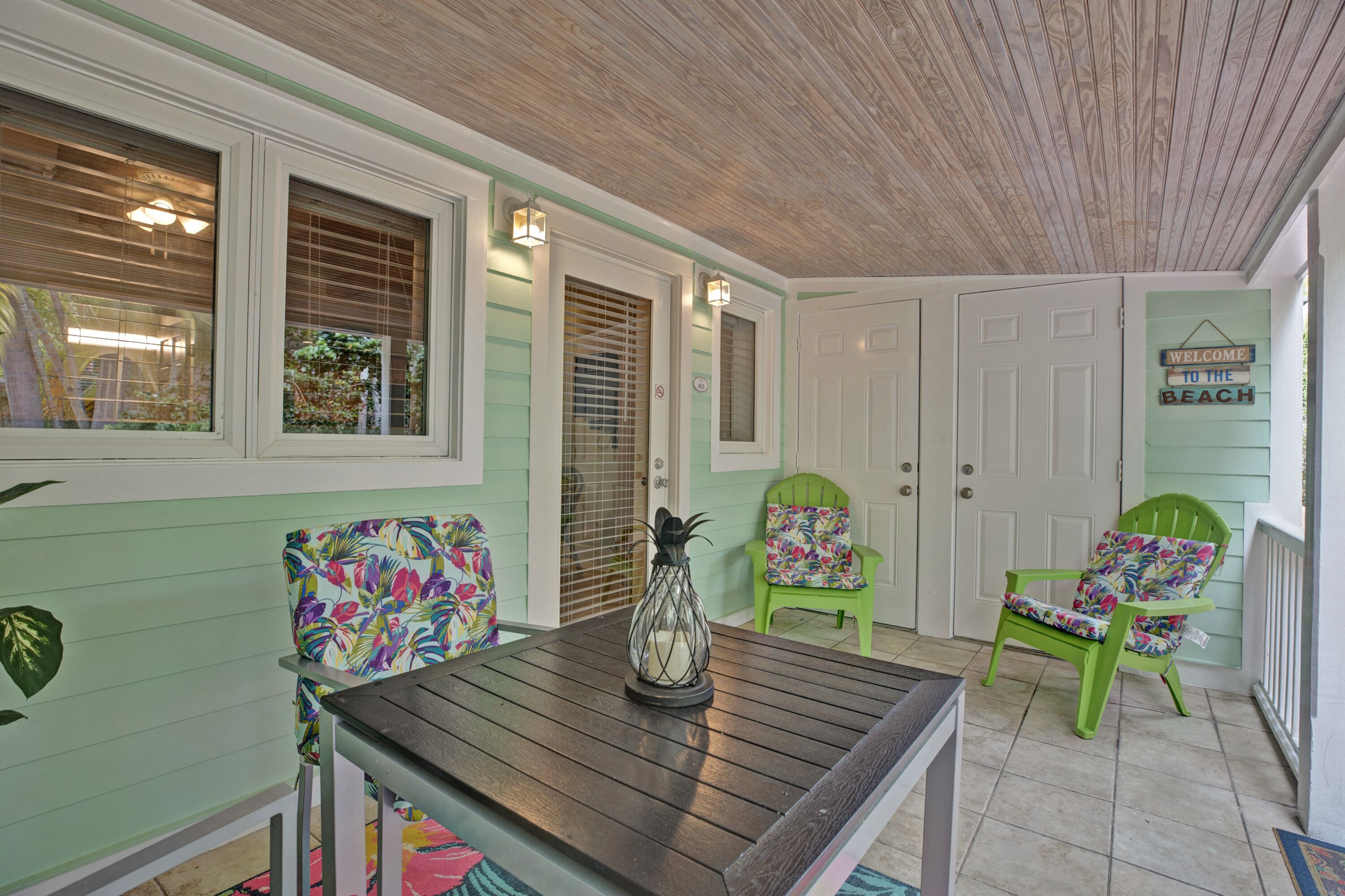 A covered porch with a wooden table, two green chairs with floral cushions, and a sign that reads "WELCOME TO THE BEACH".