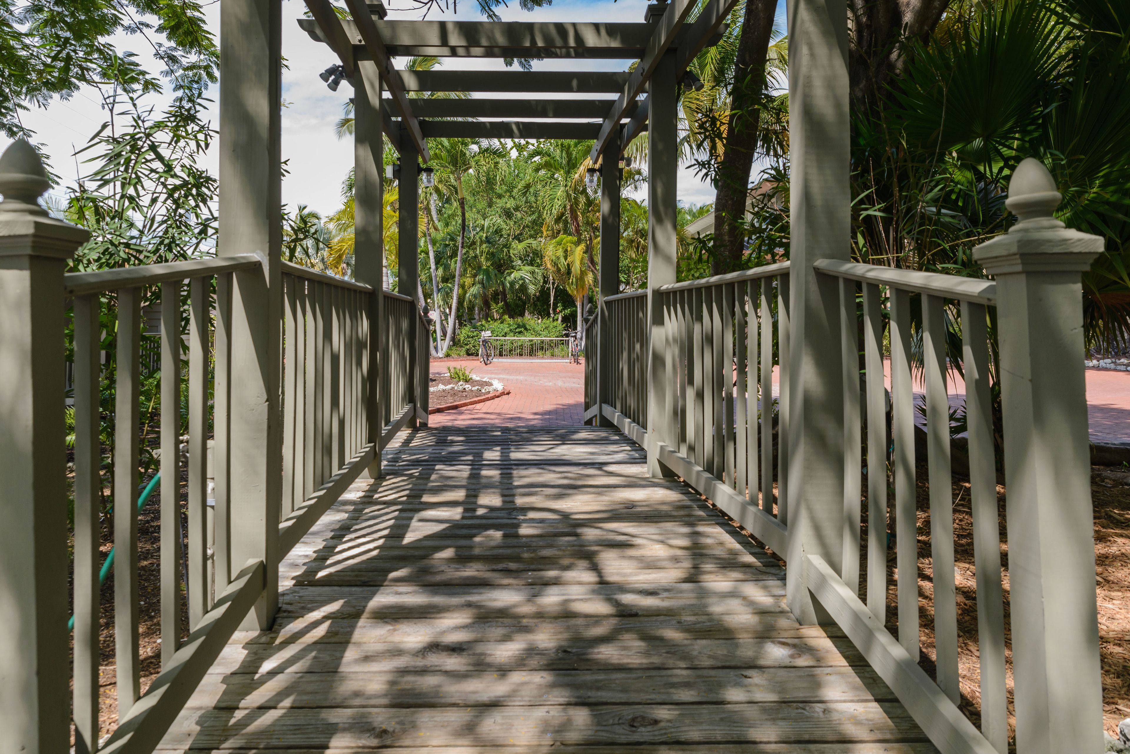 A wooden pathway leading to a covered area with a pergola structure, surrounded by greenery and palm trees.