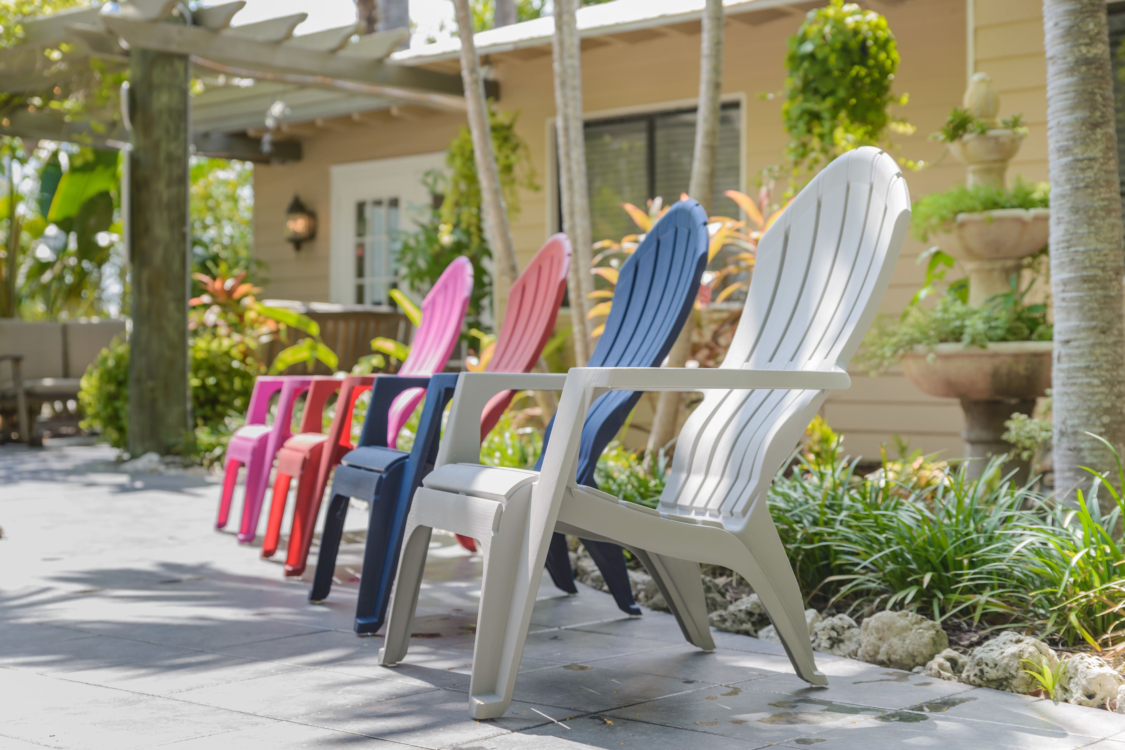 A row of colorful Adirondack chairs lined up on a patio with a pergola overhead and potted plants around.