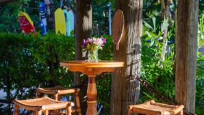 A wooden table with a vase of flowers, surrounded by wicker chairs, set against a backdrop of trees and a wooden structure.