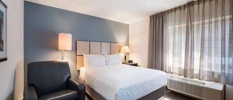 Una habitación de hotel con una cama, una silla, una mesita de noche, una lámpara y un ventanal con cortinas.