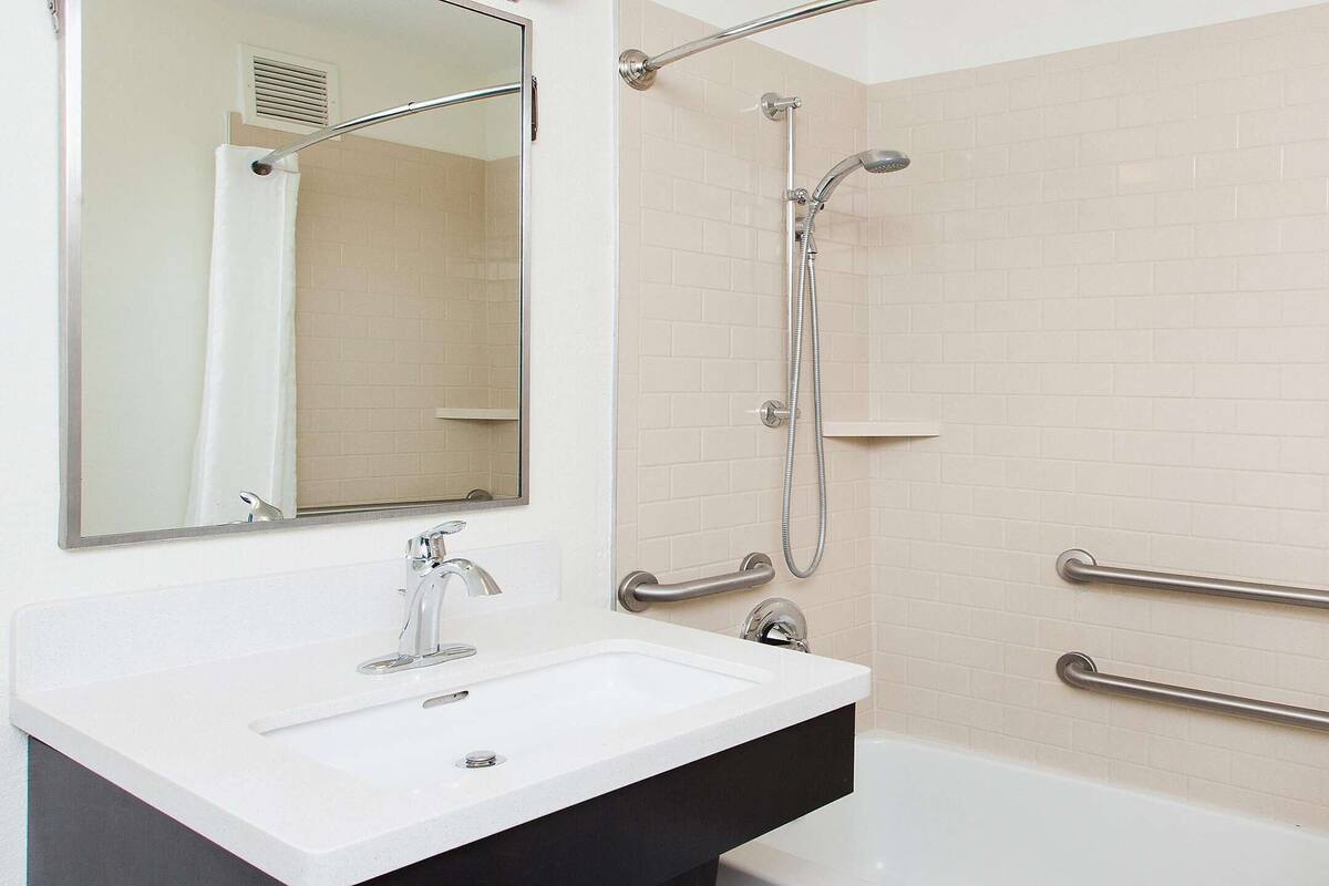 A bathroom with a white sink, a mirror, a shower with a handheld showerhead, and grab bars on the wall.