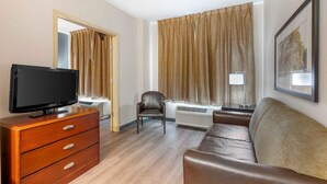 Habitación de hotel con televisión de pantalla plana sobre un tocador de madera, sofá de cuero, una silla y un ventanal con cortinas.