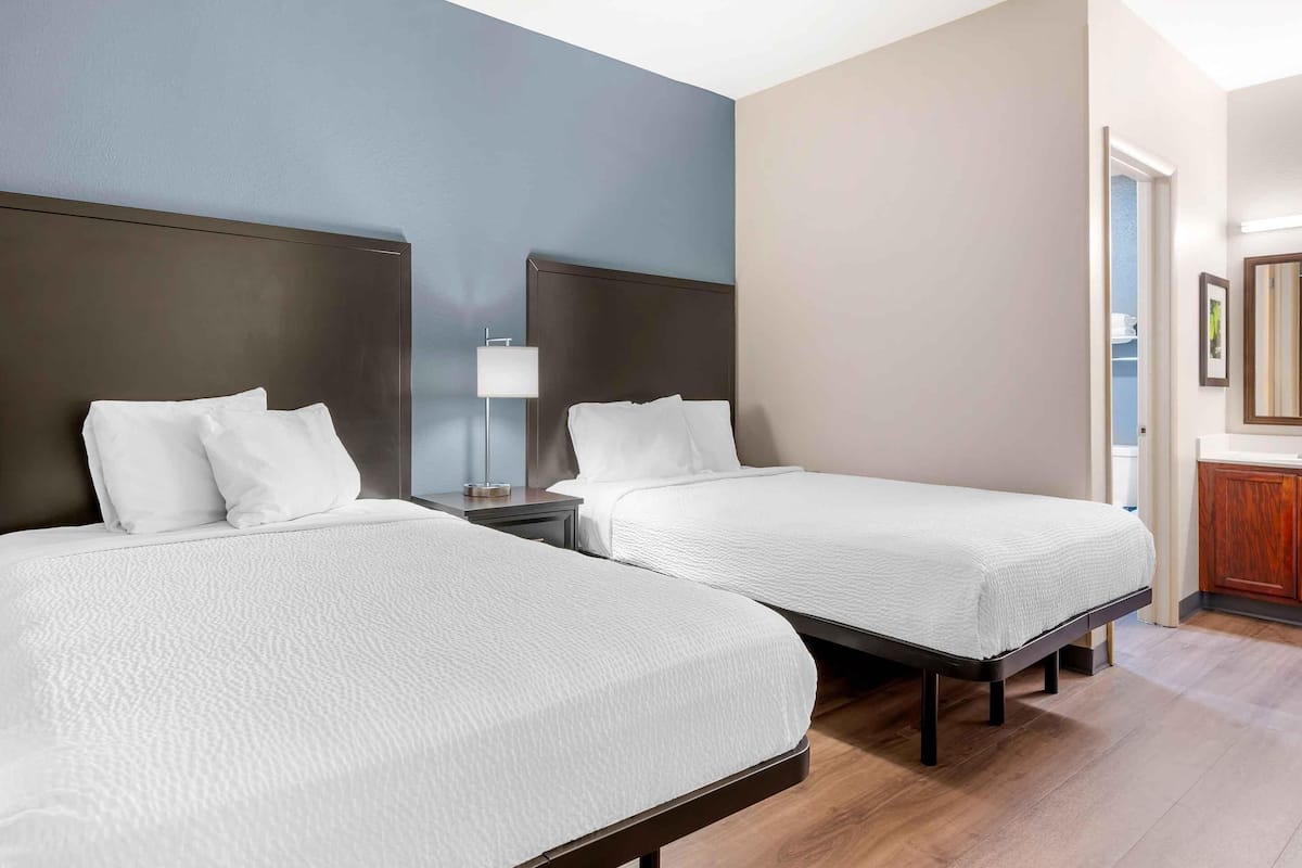 Una camera d'albergo con due letti, un comodino con una lampada, pavimento in legno e bagno visibile attraverso una porta aperta.