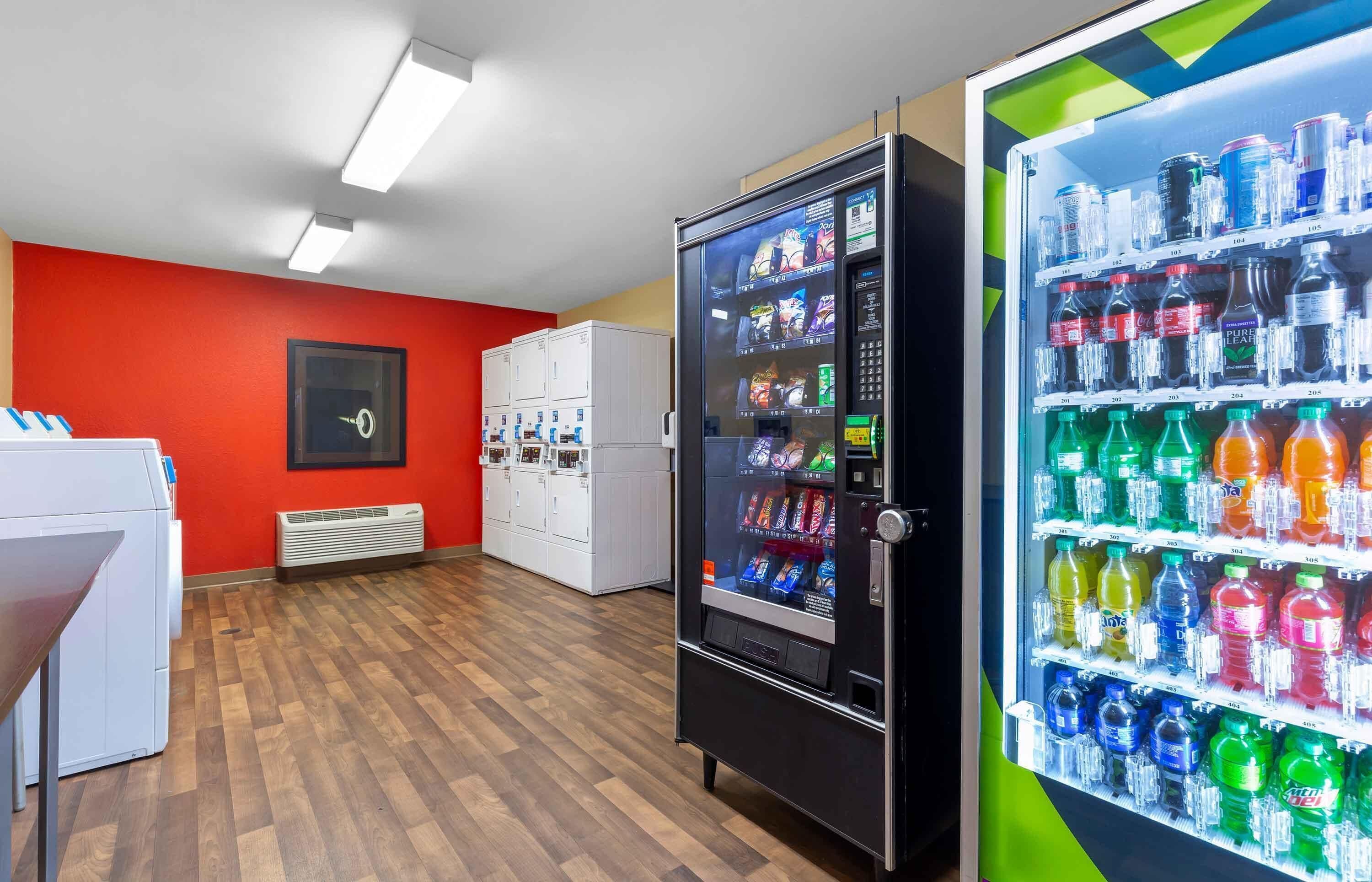 Ein Raum mit einem Automaten, gefüllt mit verschiedenen Getränken und Snacks, einer weißen Waschmaschine und einer roten Wand.
