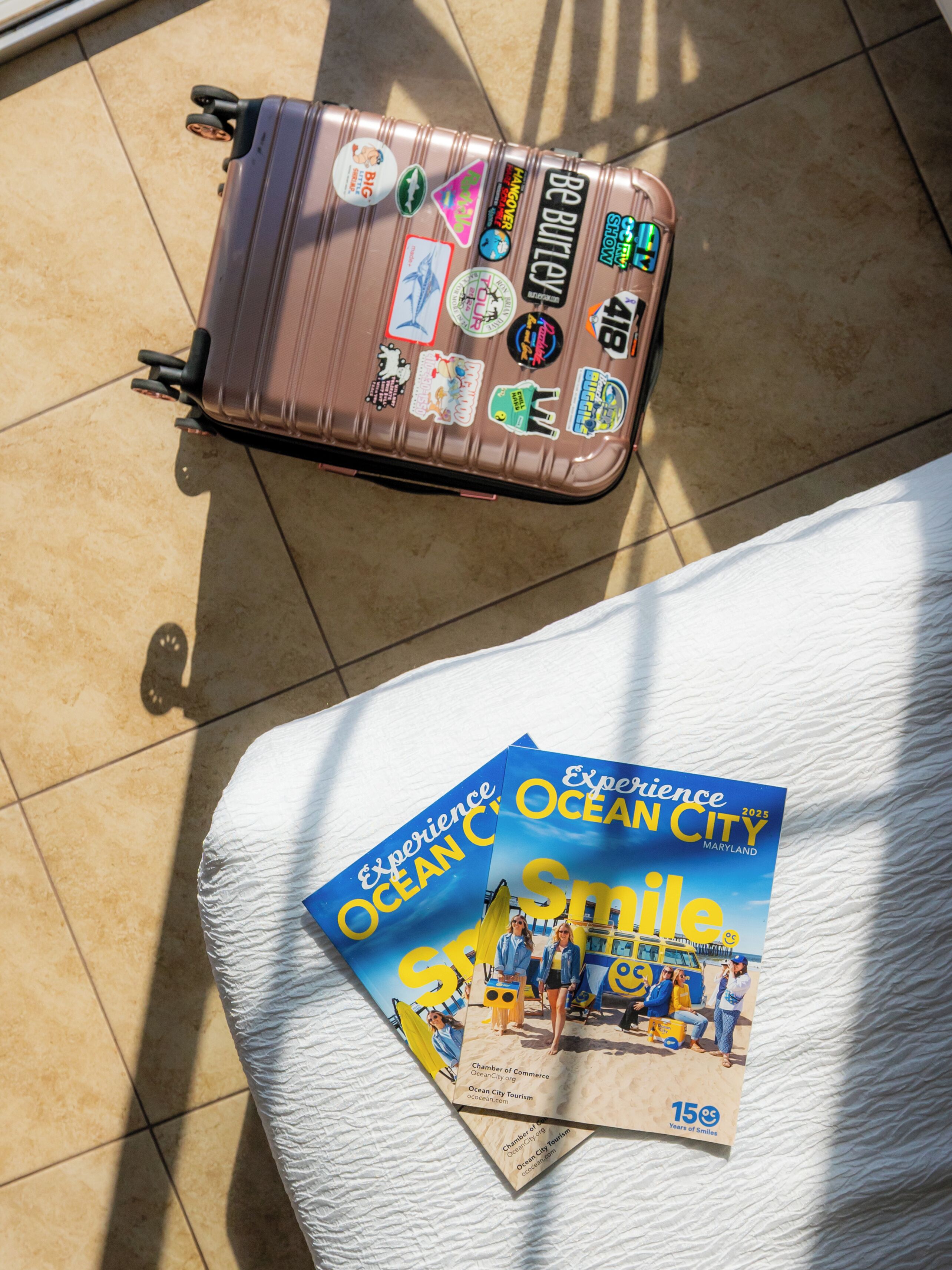 A suitcase with travel stickers and a brochure titled "Experience Ocean City 2025" on a bed.
