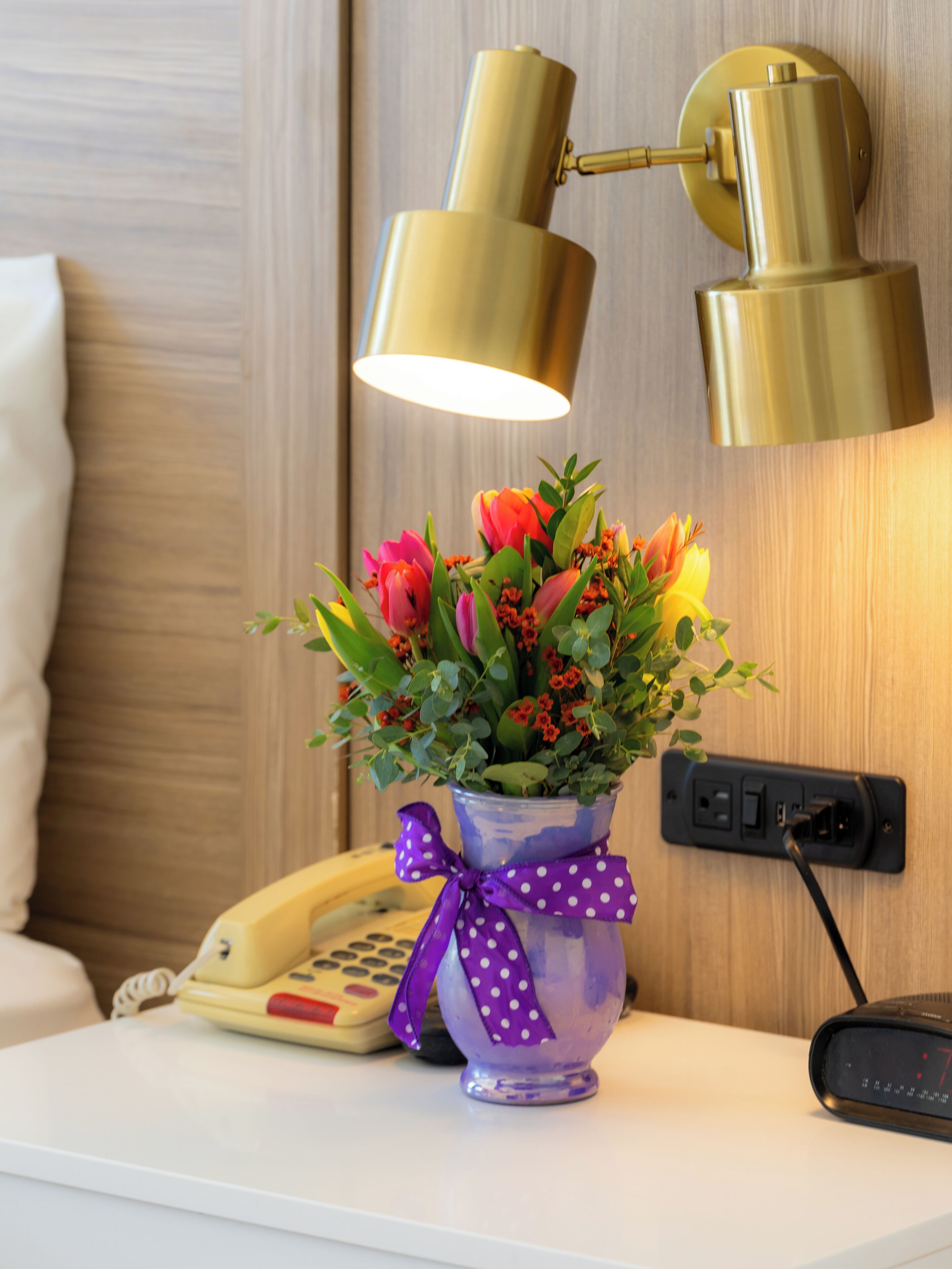 A bedside table with a vase of flowers, a telephone, and a lamp.
