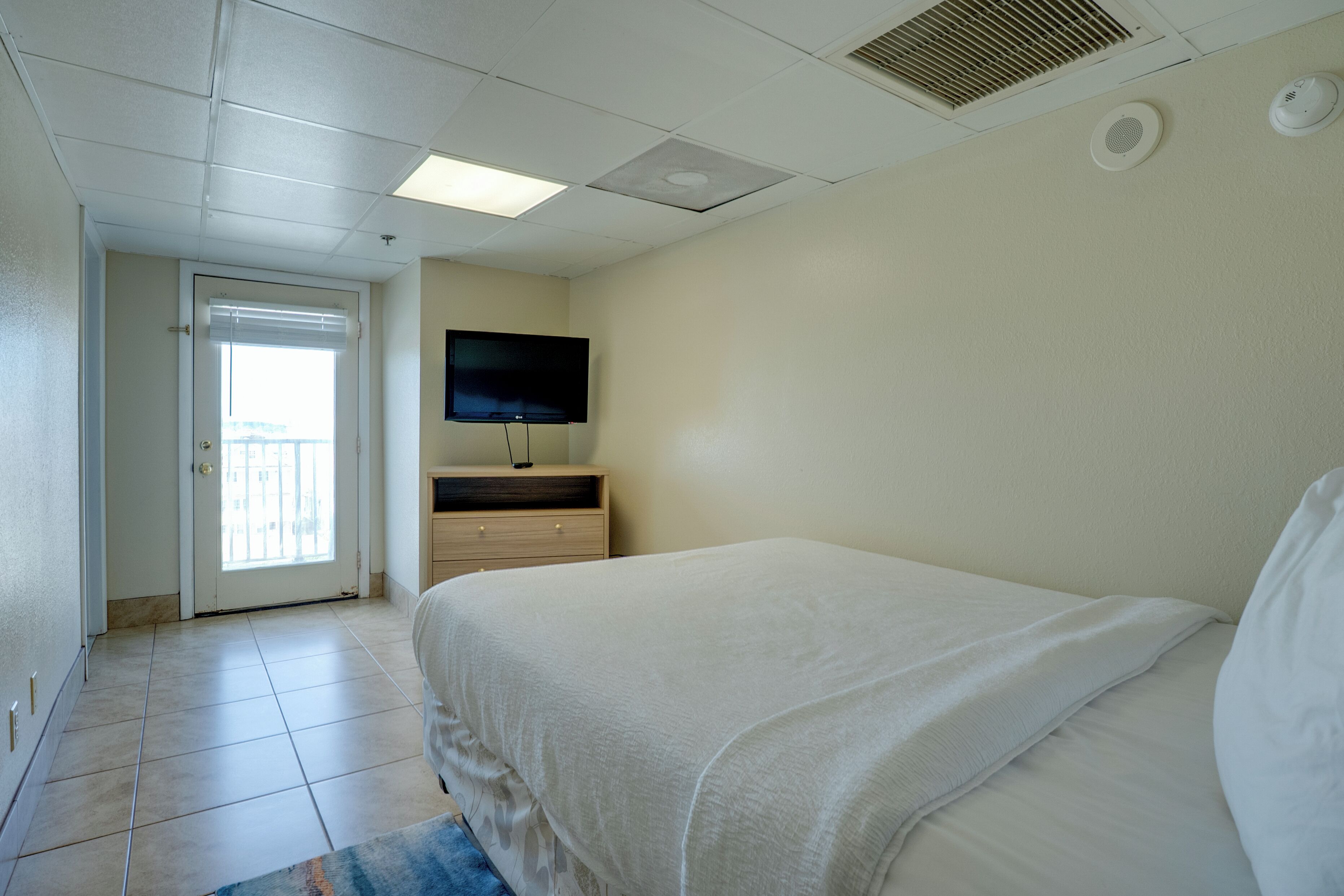 A hotel room with a bed, a TV mounted on the wall, a door leading to a balcony, and a ceiling vent.