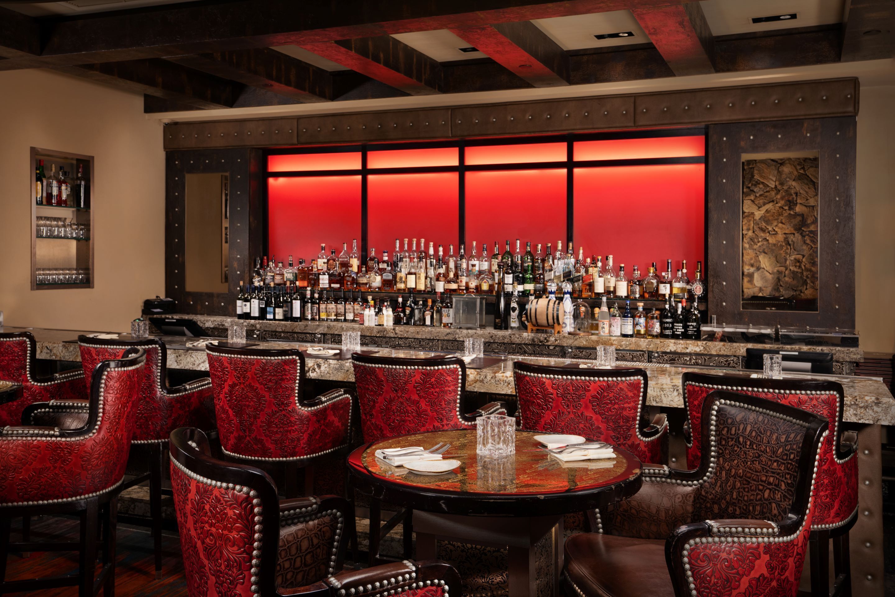 A bar area with a red-lit backdrop, multiple liquor bottles, and red upholstered chairs.