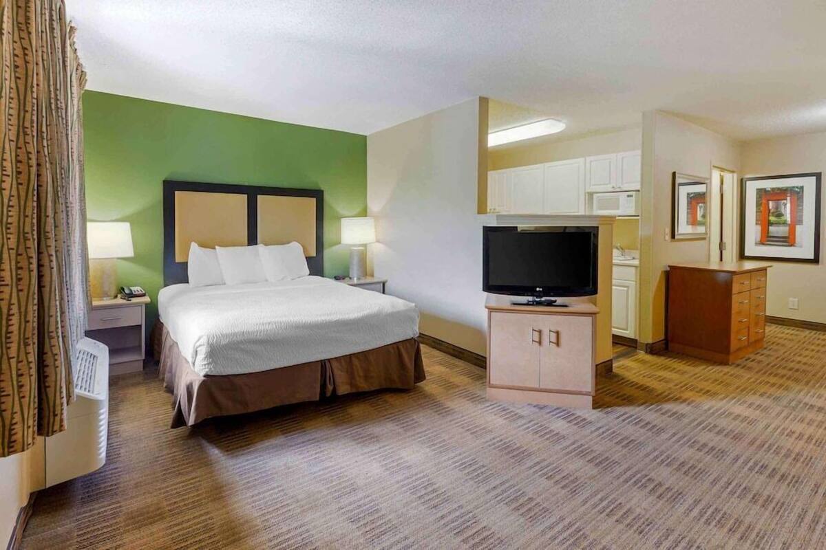 A hotel room with a bed, bedside lamps, a TV, a small cabinet, and a microwave in the kitchenette.