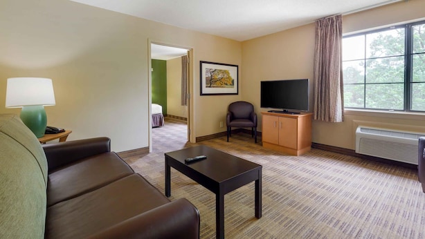 A hotel room with a sofa, a small table, a television, a cabinet, and a window with curtains. - Extended Stay America Select Suites Raleigh RTP 4610 Miami B (Durham)