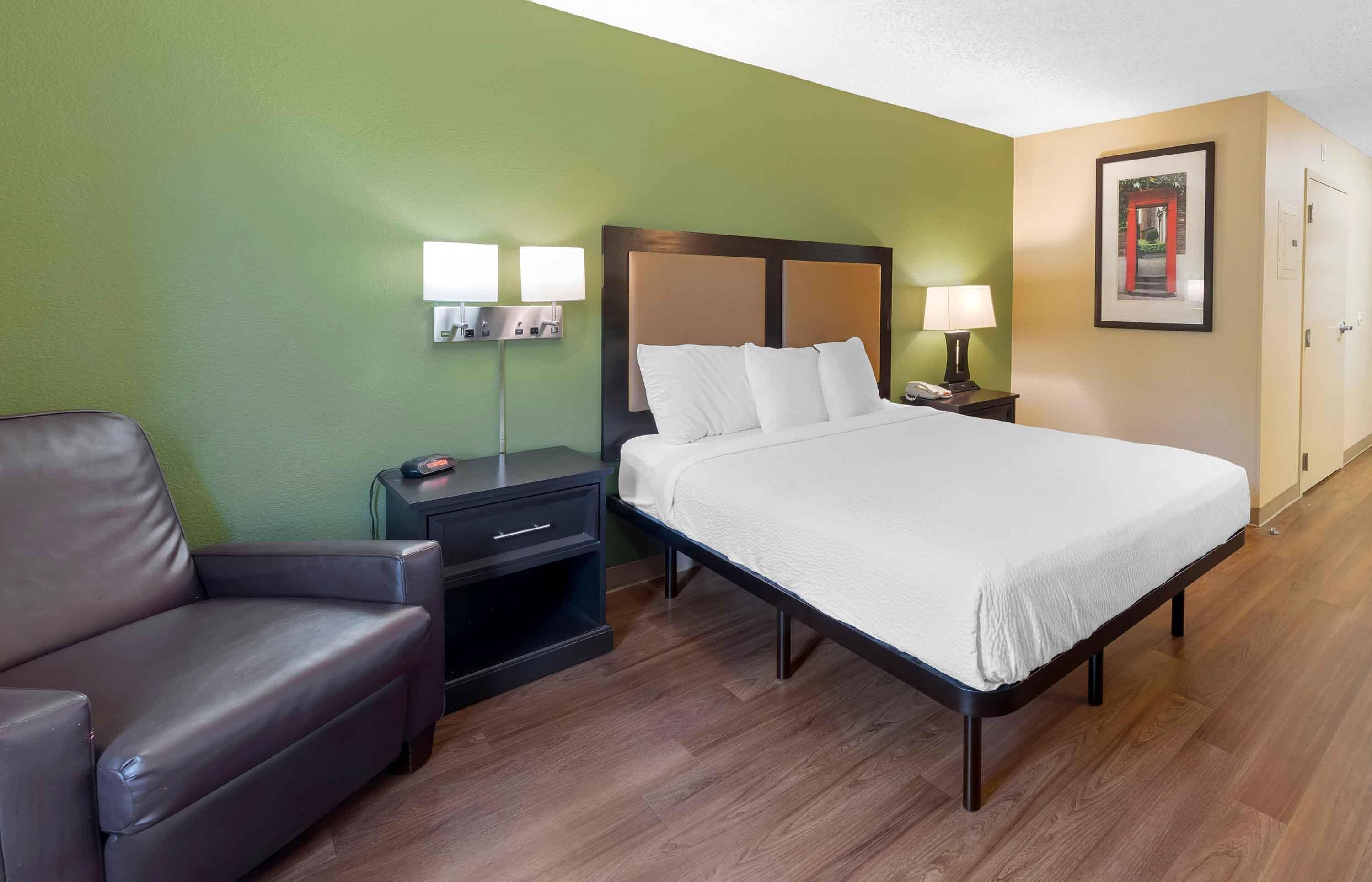 A hotel room with a bed, a nightstand, a lamp, a chair, and a framed picture on the wall.