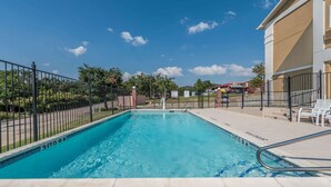 Una piscina con agua cristalina azul, rodeada por una cerca y un edificio de fachada color beige.