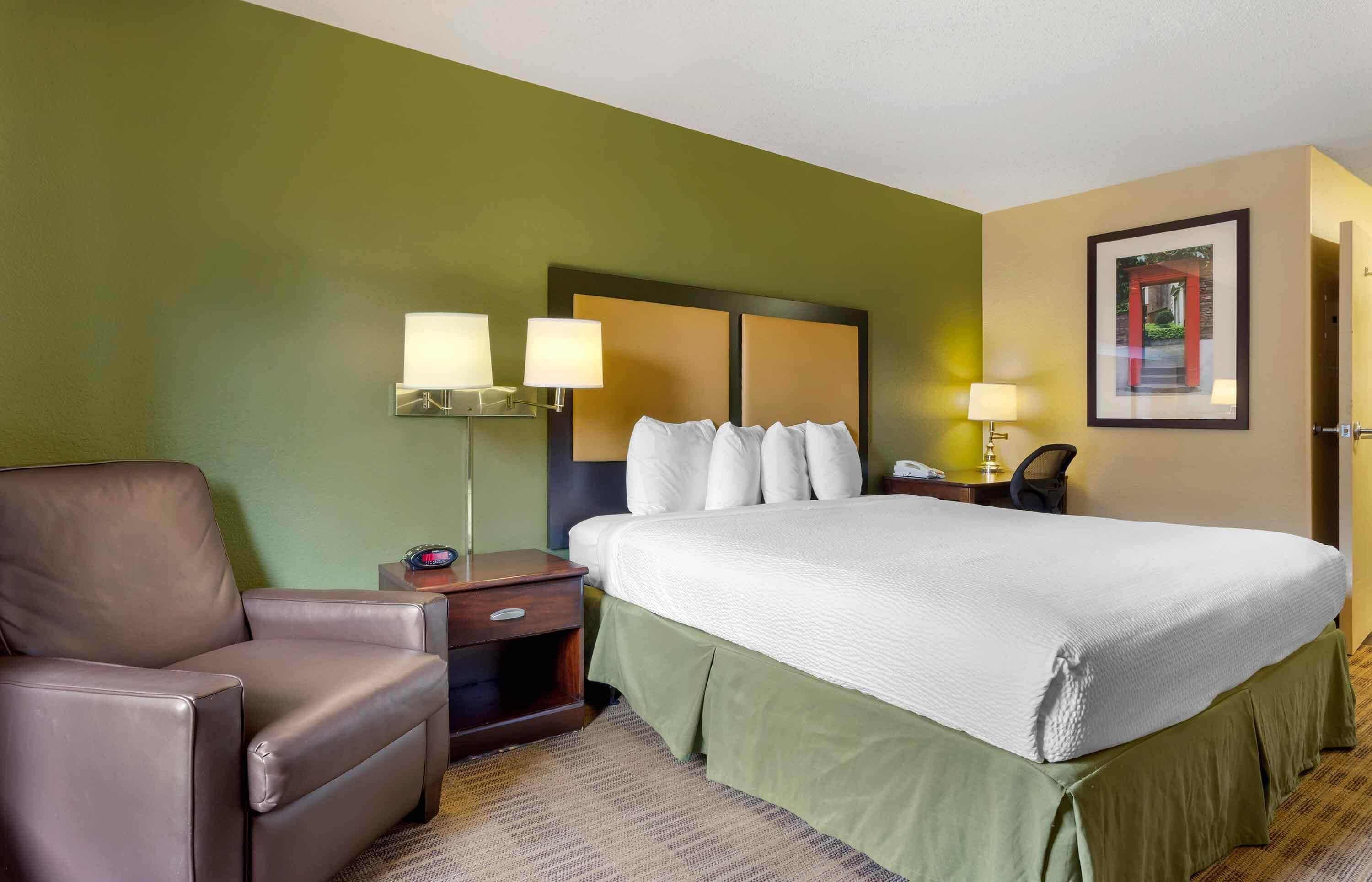 A hotel room with a large bed, a chair, a nightstand with lamps, and a framed picture on the wall.