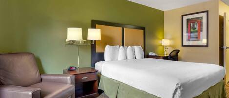 A hotel room with a large bed, a chair, a nightstand with lamps, and a framed picture on the wall.