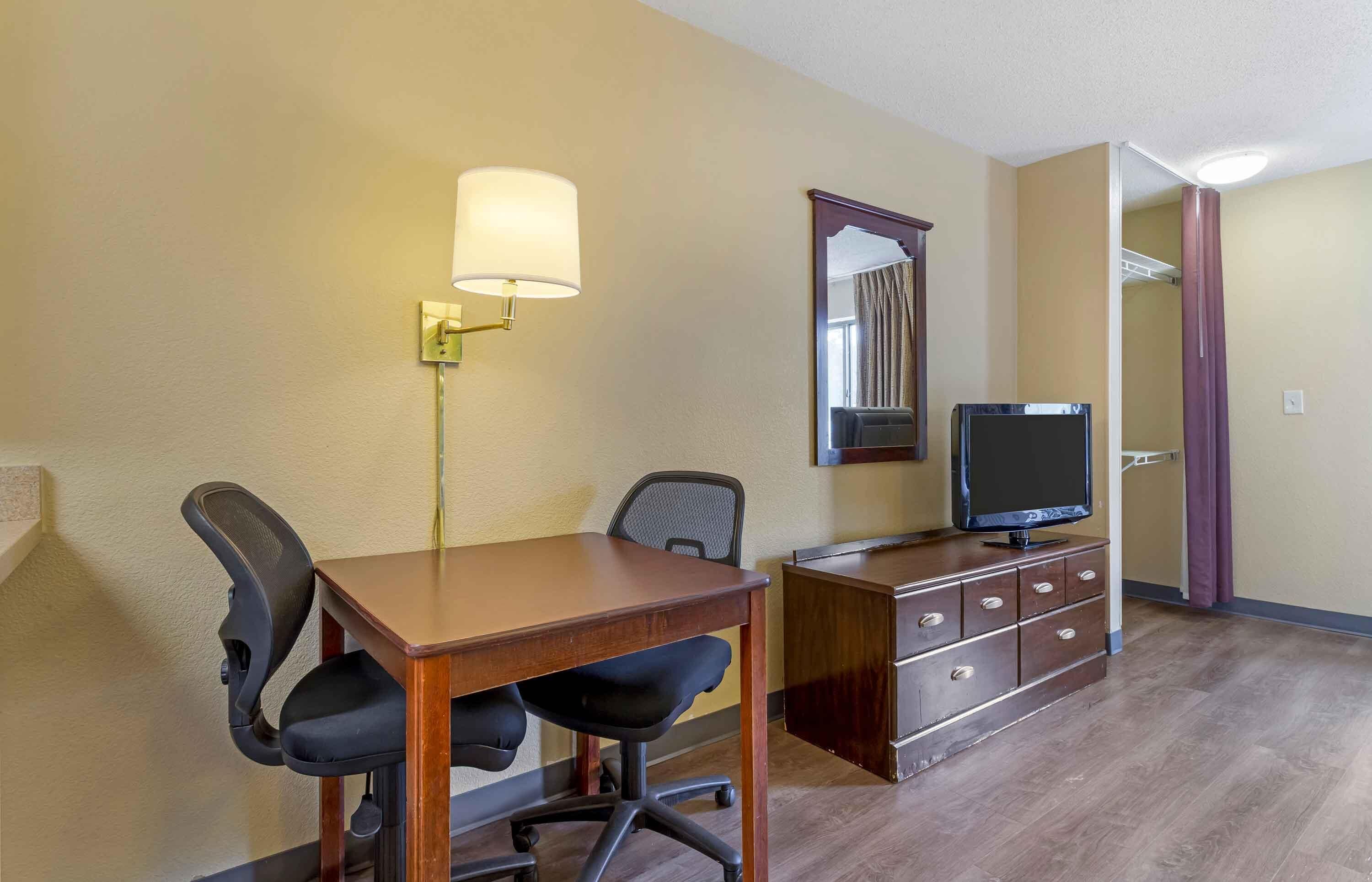 A hotel room with a wooden desk, two black office chairs, a television on a wooden cabinet, a wall-mounted lamp, and a mirror.