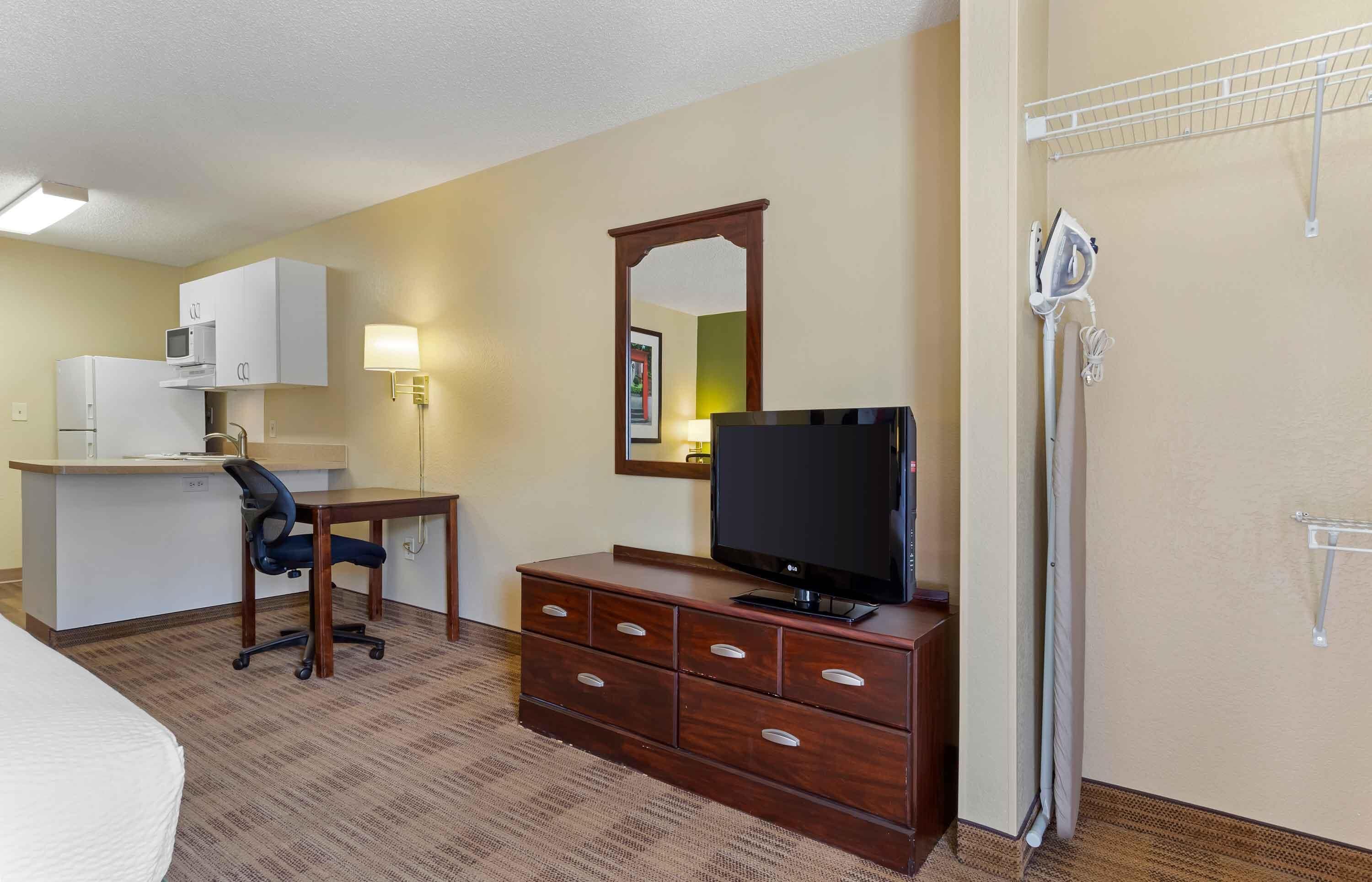 A hotel room with a kitchenette, a desk with a chair, a TV on a dresser, and a shower area.